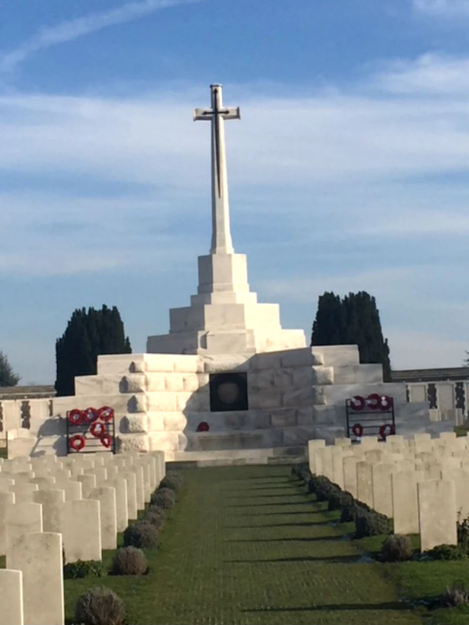 A cemetery with rows of white gravestones leading to a large stone memorial cross, adorned with wreaths and surrounded by trees.