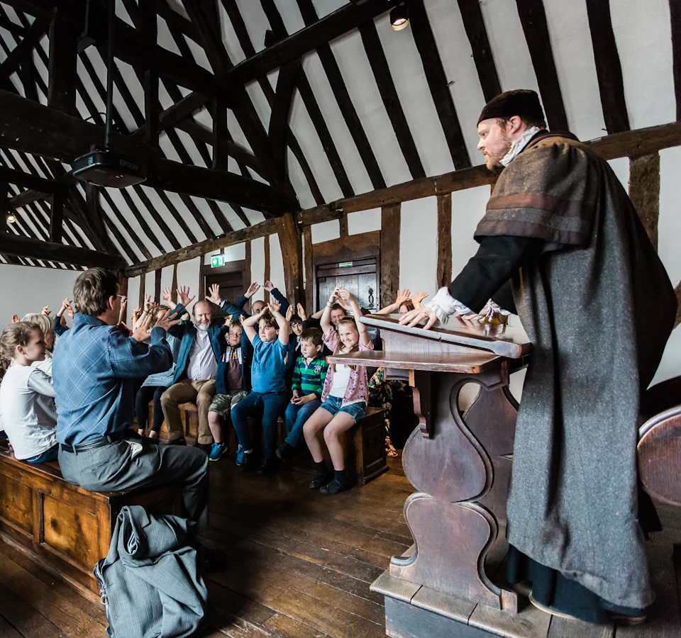 Man in period costume lecturing children seated on benches in a room with wooden beams. Children have raised hands.