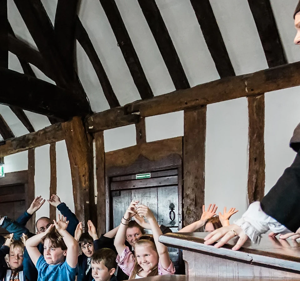 Man in period costume lecturing children seated on benches in a room with wooden beams. Children have raised hands.