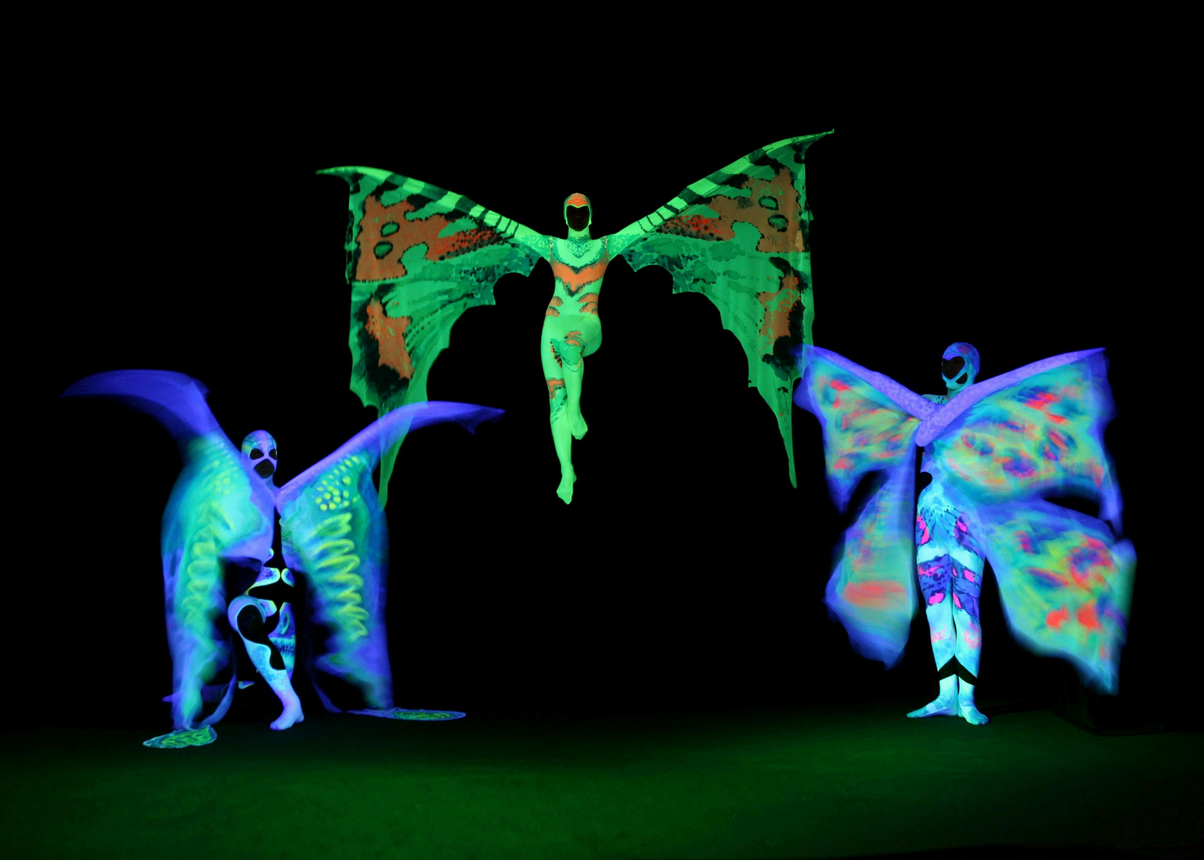 Three performers in glowing butterfly costumes with vivid, neon colors against a black background, one performer is airborne.