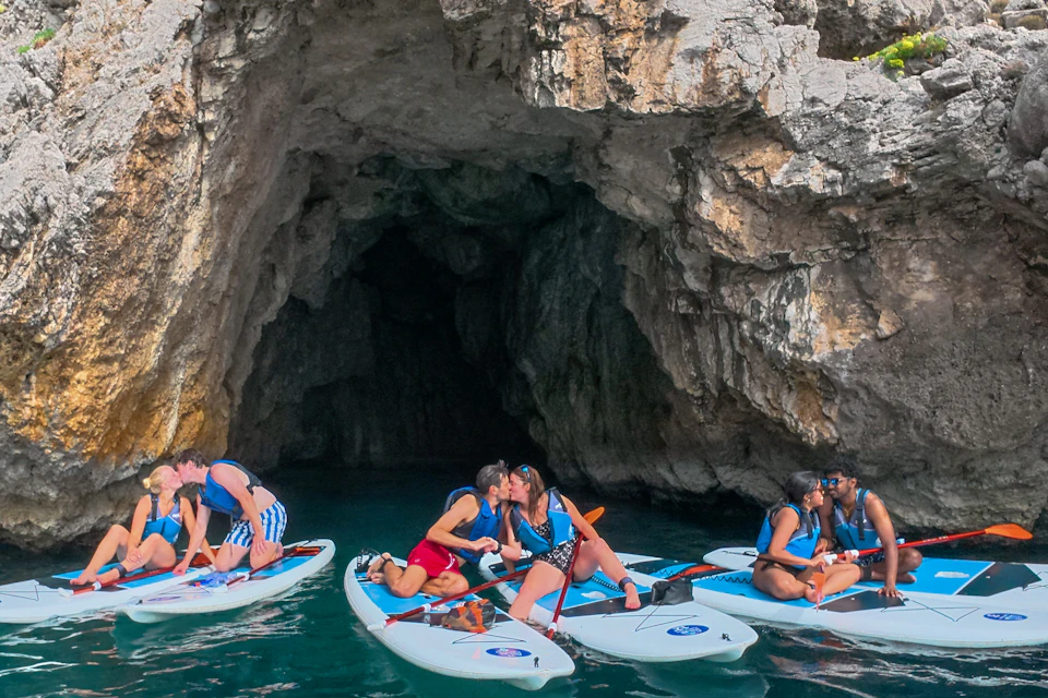 A group of people on paddleboards near a rocky cave, with some couples kissing and others enjoying the activity.