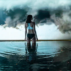 En person i baddräkt står på knä i vatten och står inför mörka stormoln i horisonten.