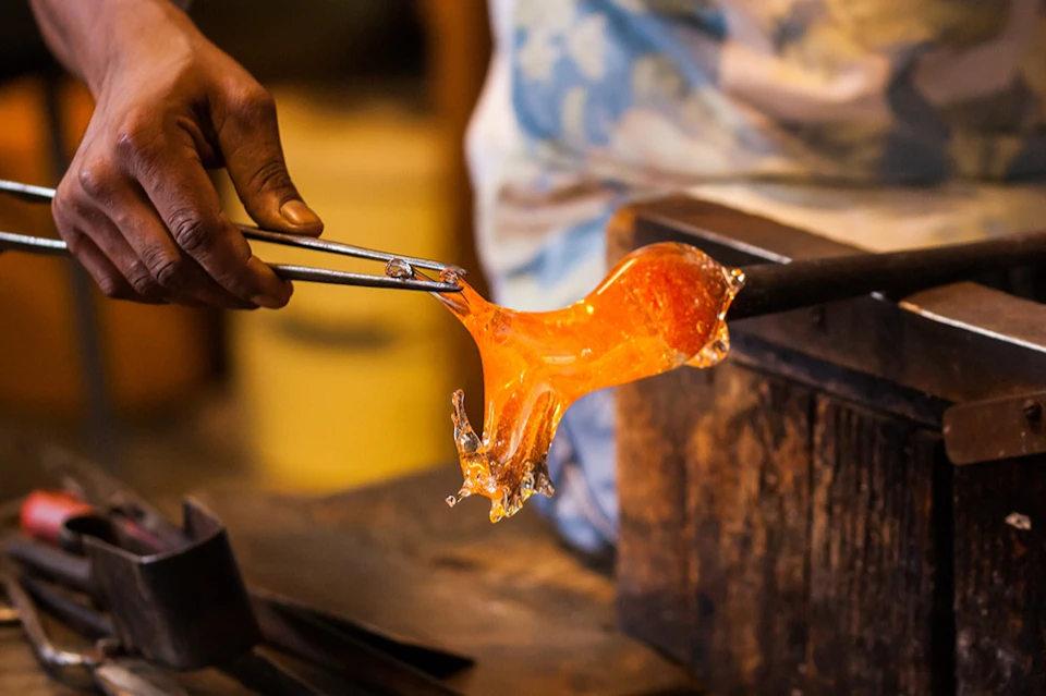 A person shaping molten glass with tongs in a workshop setting.