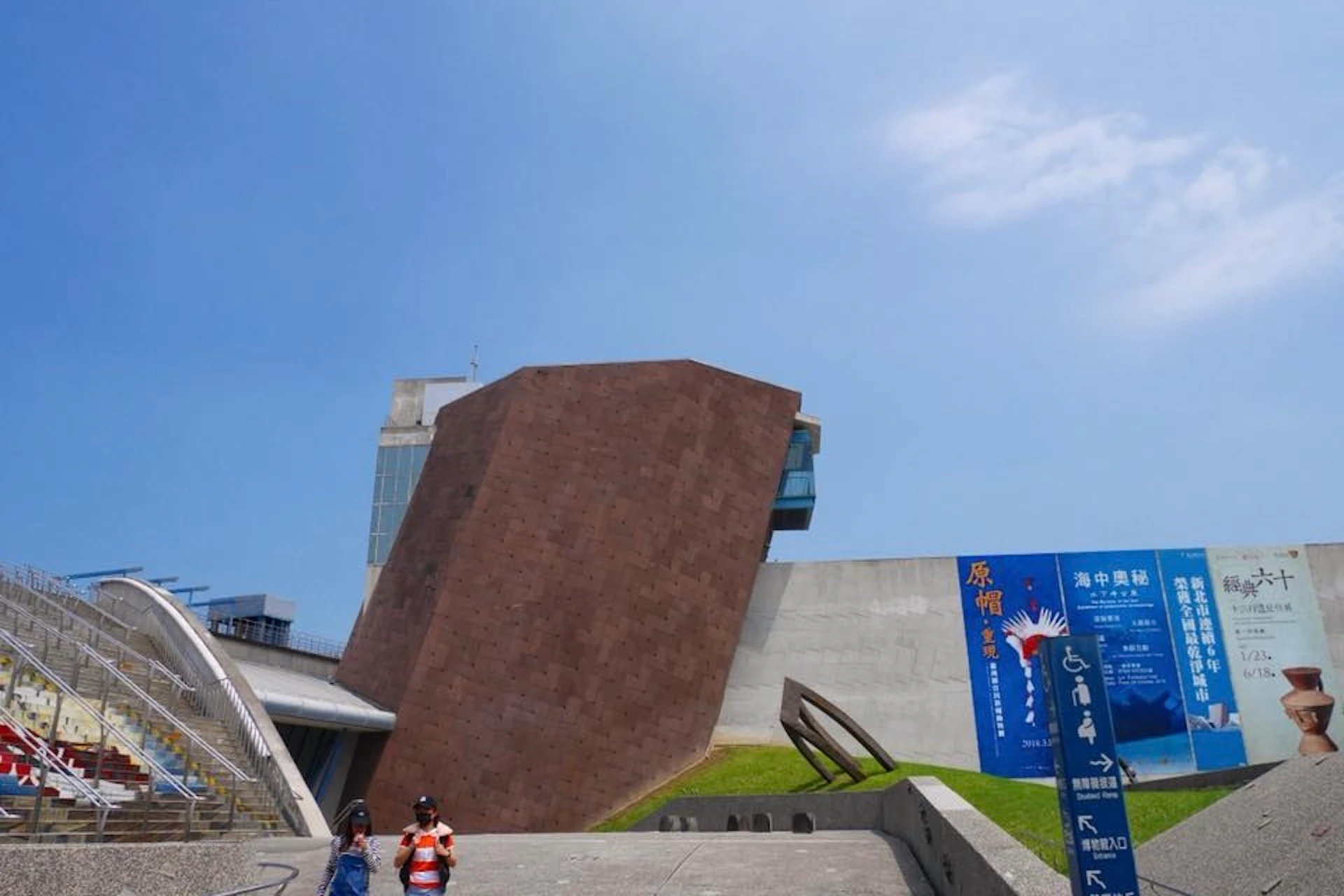 A modern building with an angular brown structure and adjacent white walls, under a clear blue sky. Two people walk in the foreground.