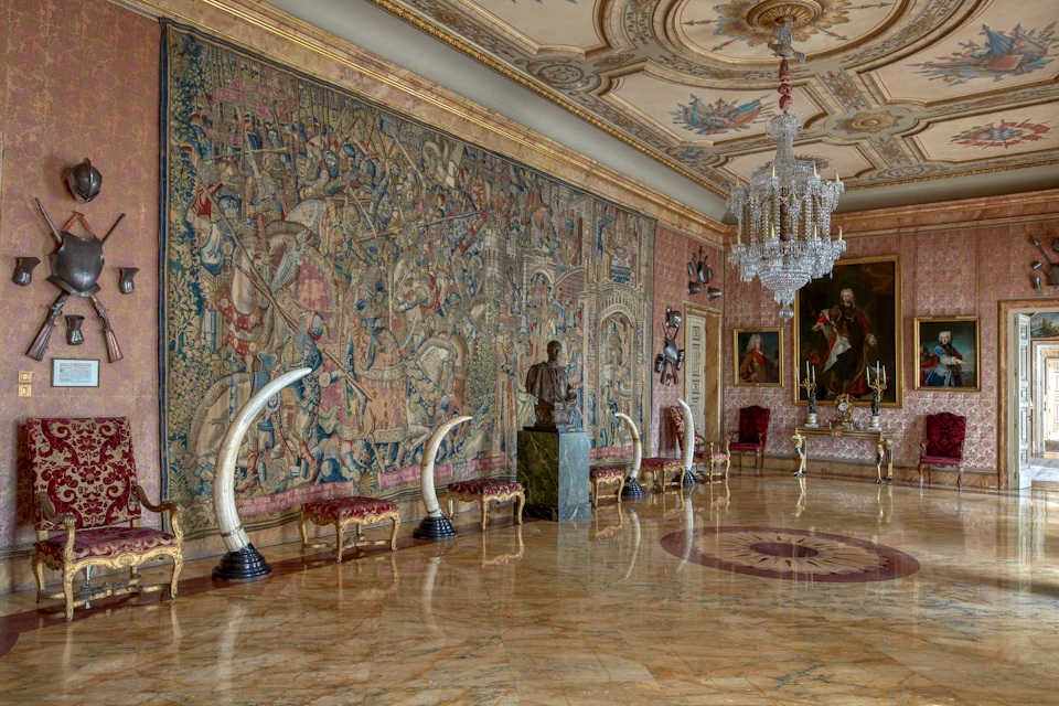 Ornate room with a large wall tapestry, marble floors, a chandelier, bust sculptures, and antique furniture including chairs and benches.