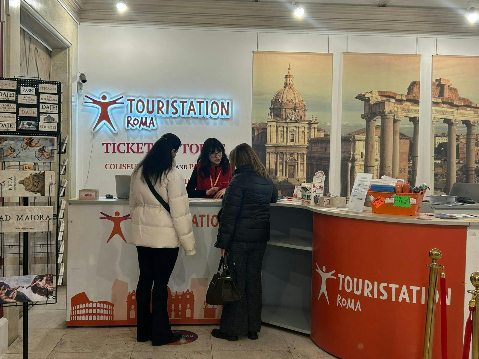 Tre persone presso un banco di informazioni turistiche con l'etichetta "Touristation Roma", con espositori promozionali e decorazioni di architettura romana.