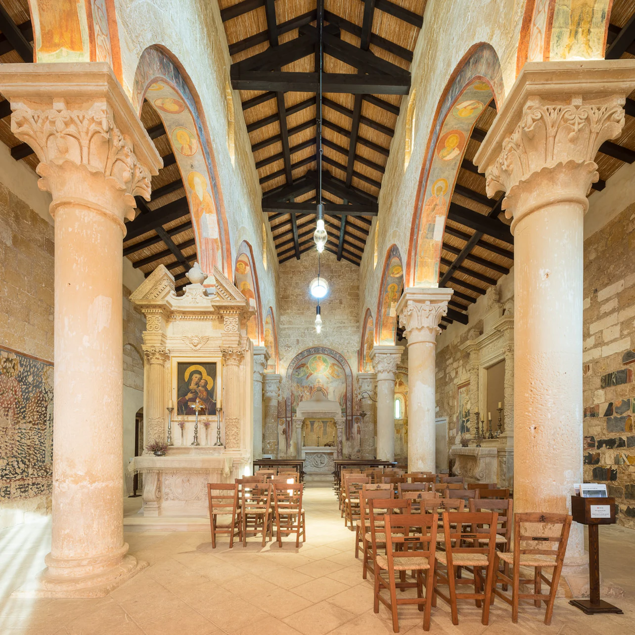 An interior of a historic church with wooden ceiling, stone columns, frescoed arches, icon paintings, and wooden chairs arranged in rows.