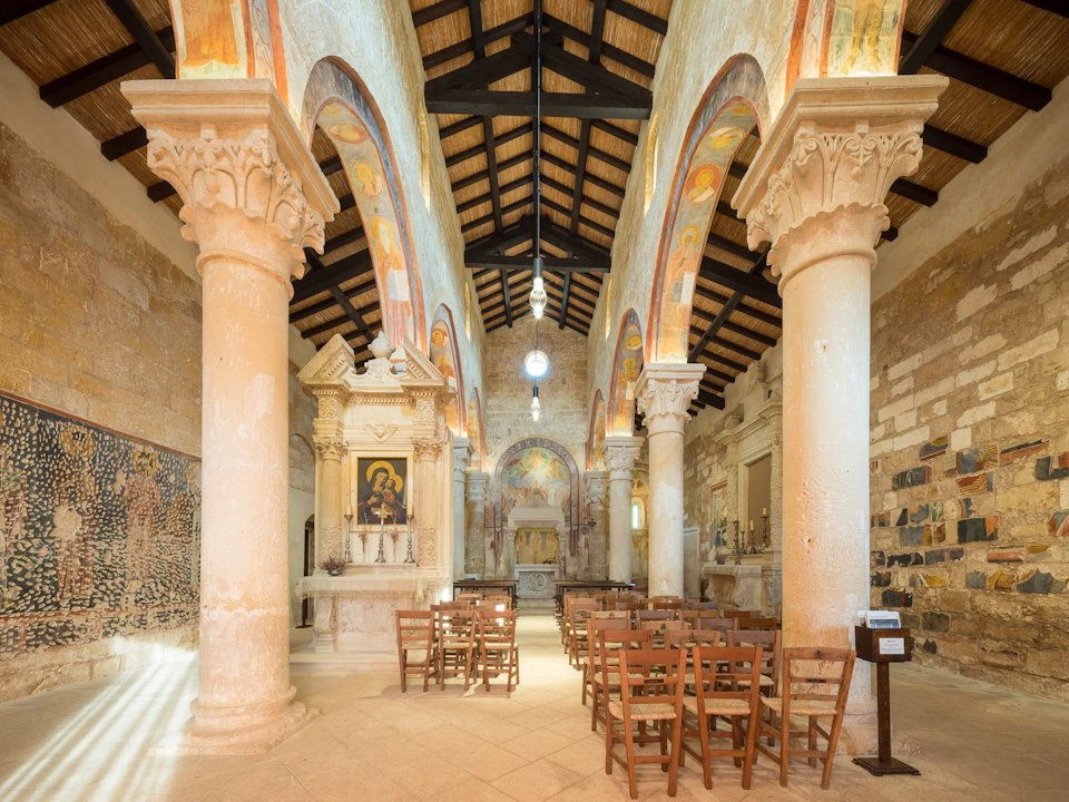An interior of a historic church with wooden ceiling, stone columns, frescoed arches, icon paintings, and wooden chairs arranged in rows.