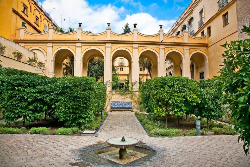 A courtyard with a fountain, manicured bushes, and an arched colonnade under a blue sky.