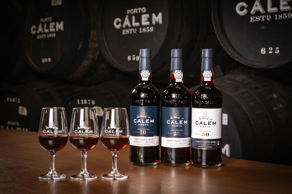 Three bottles of Calem Porto wine with glasses of wine on a table, barrels in the background.