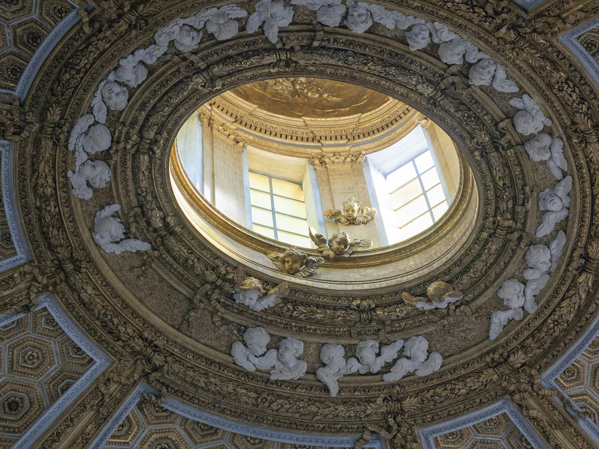 Plafond circulaire orné d'accents décoratifs or et blanc, avec des visages et des anges sculptés autour d'une lucarne centrale.