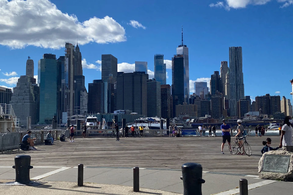 Skyline of tall buildings under a blue sky with clouds, people walking and biking on a boardwalk, and boats docked by the water.
