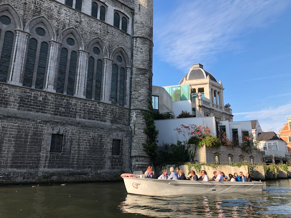 People in a small tour boat pass by historic stone buildings along a canal under a clear blue sky.