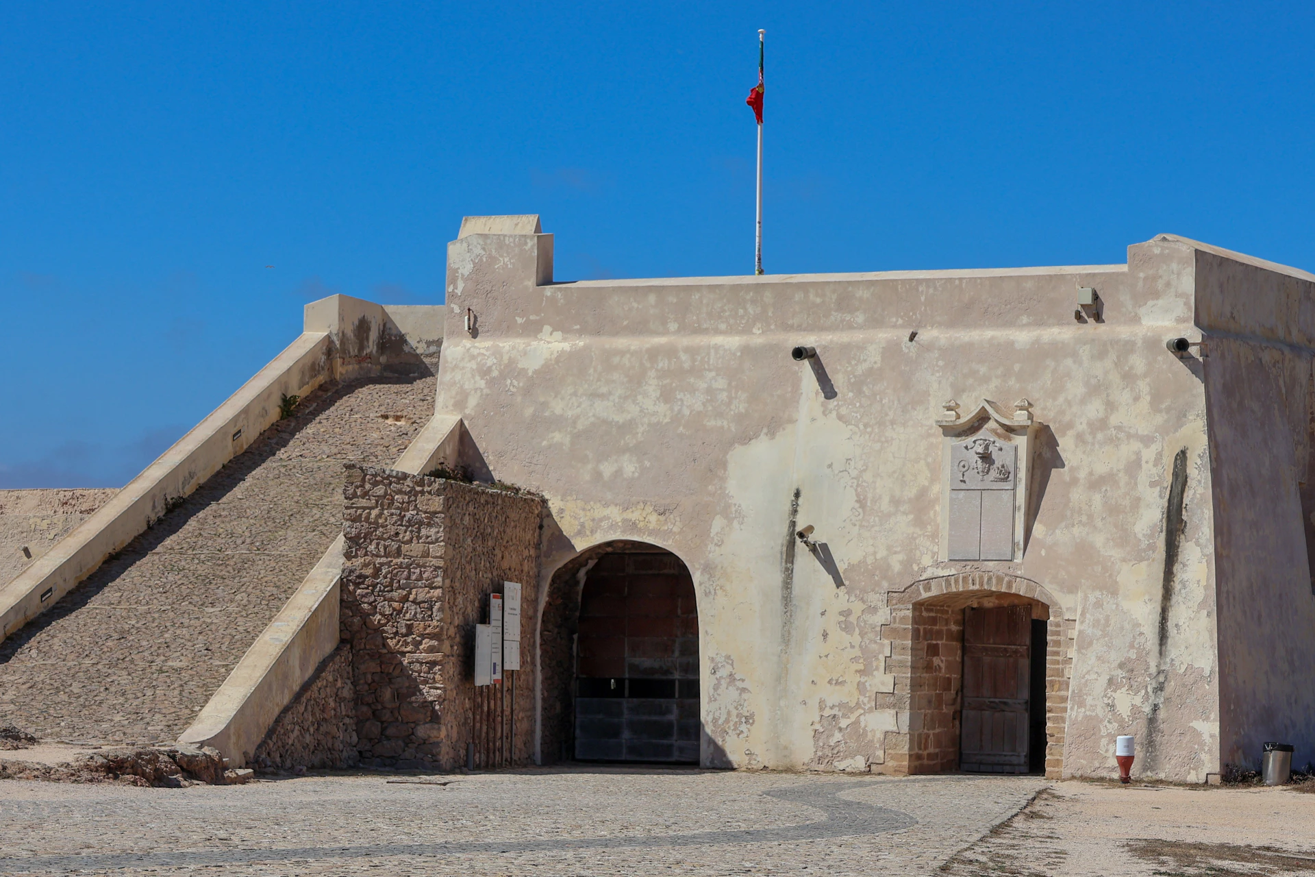 A historic fort with arched entrances, a flagpole with a red flag, and a stone wall under a clear blue sky.