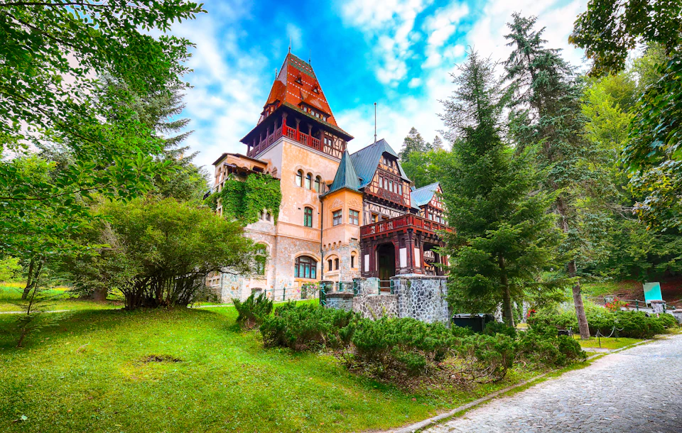 A large, ornate, multi-story building with mixed architectural styles surrounded by lush greenery under a vibrant, partly cloudy sky.