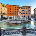 A large stone fountain with multiple statues surrounded by a small pool in a plaza, with colorful buildings in the background.