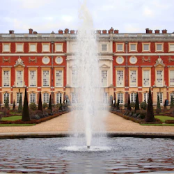 Ett historiskt palats i rött tegel med stora fönster, en central fontän på innergården, statyer, trimmade häckar och en klar himmel ovanför.