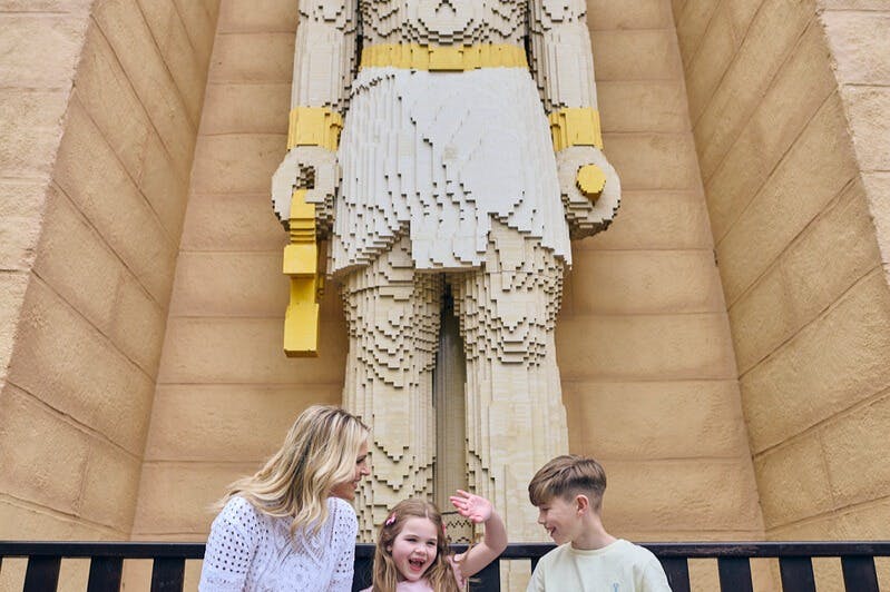 Uma grande estátua Lego de uma figura vestida de branco e amarelo está atrás de uma mulher e duas crianças que sorriem e interagem.
