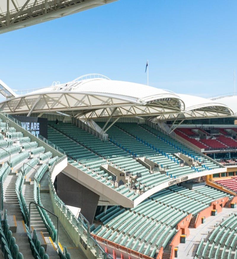 Large, empty stadium with green and red seats, white canopy roof, and a clear blue sky.