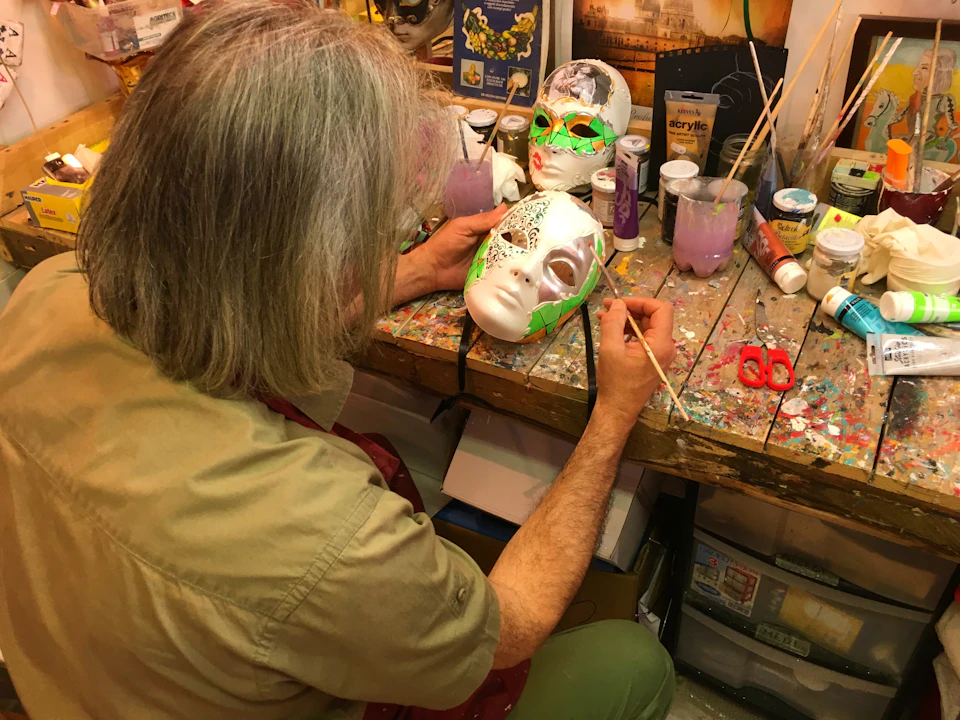 A person decorates a white mask with green and gold paint at a cluttered art workspace, with other artistic supplies around.