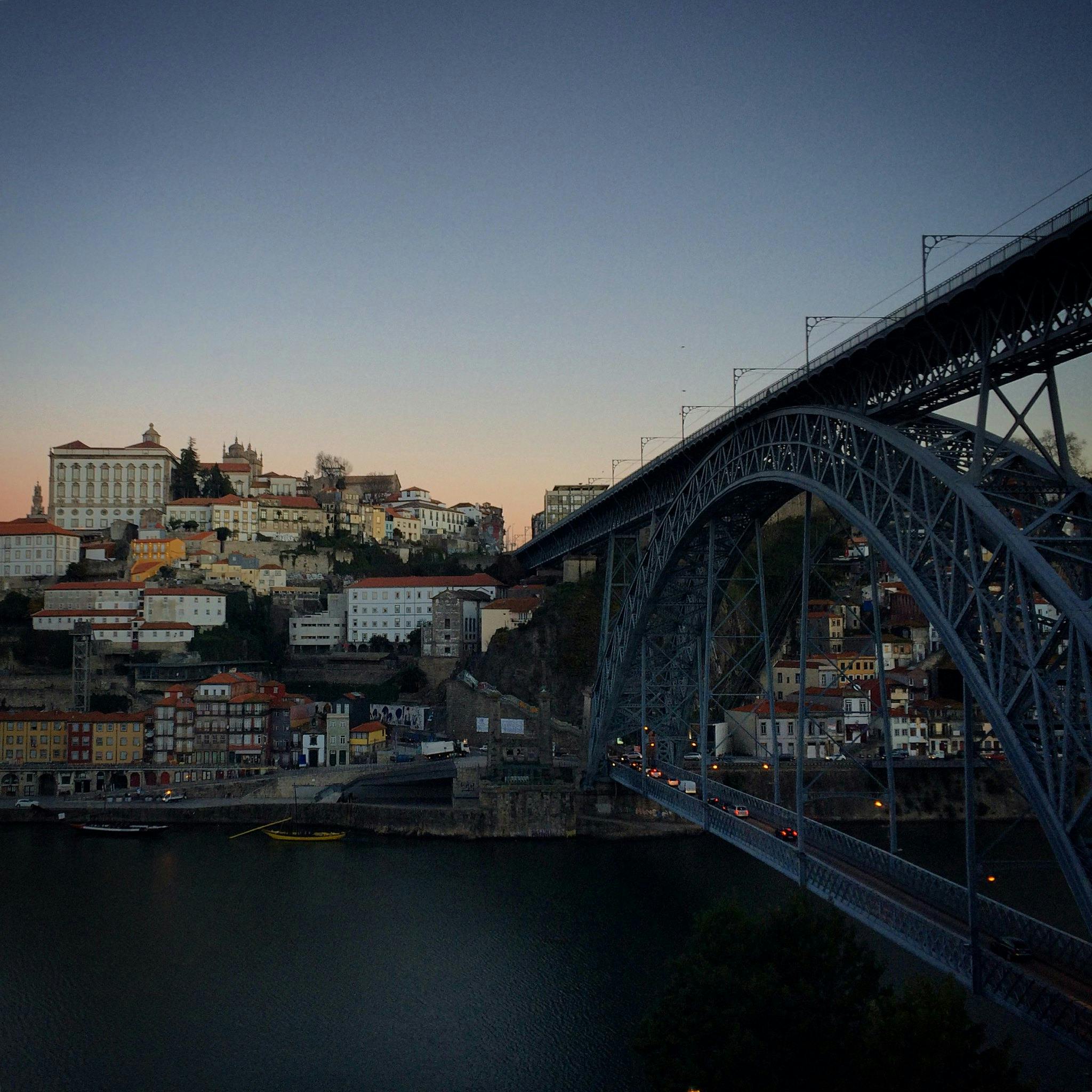 En bro sträcker sig över en flod med en stad av klusterhus och byggnader på en sluttning i skymningen.