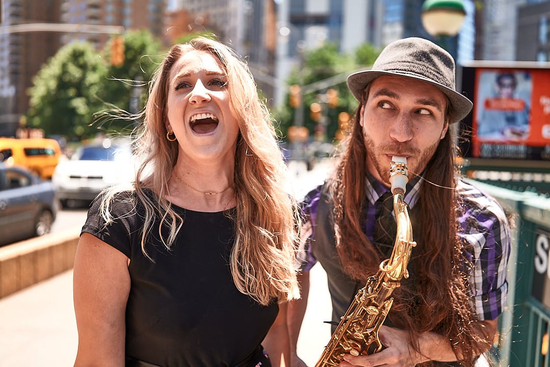 Δύο άνθρωποι έξω, μια γυναίκα που γελάει και ένας άντρας που παίζει σαξόφωνο. Αστικό φόντο με θολά κτίρια και πράσινο.