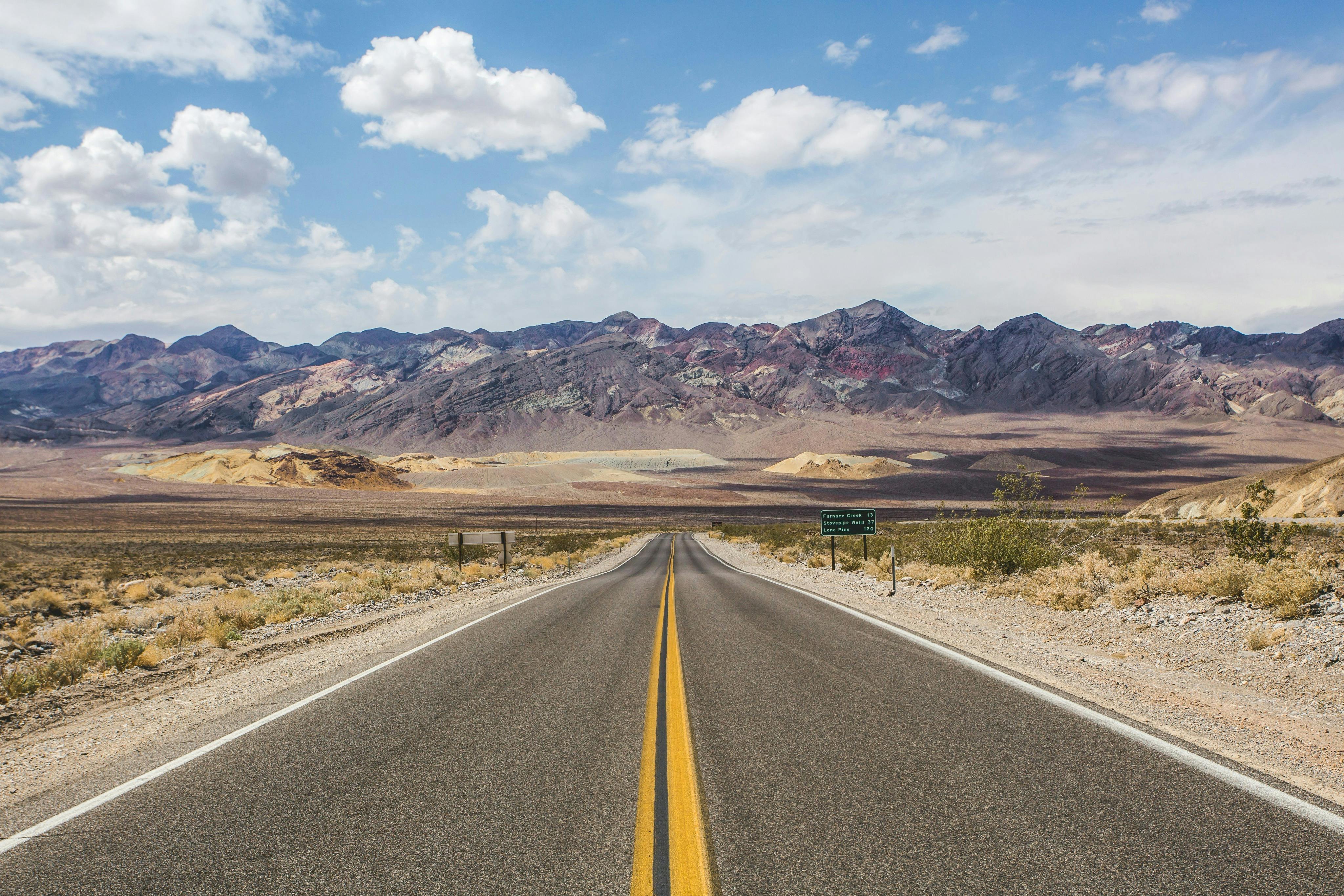 A straight road leading through a desert landscape towards distant colorful mountains under a partly cloudy sky.