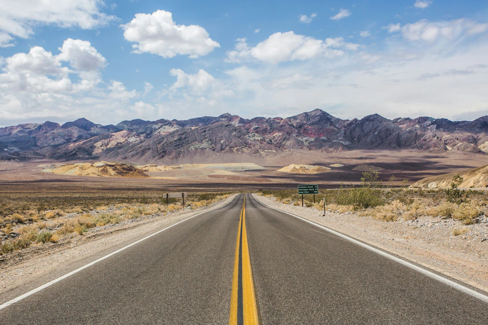 A straight road leading through a desert landscape towards distant colorful mountains under a partly cloudy sky.