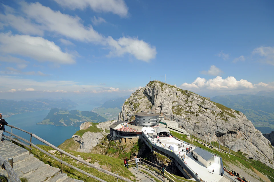 Mountain-top building with observation deck, surrounded by rugged cliffs and paths, under a clear blue sky with scattered clouds.