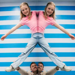 En ung flicka i rosa och jeans med en optisk illusion som får hennes kropp att se speglad ut, i ett rum med blårandiga väggar.