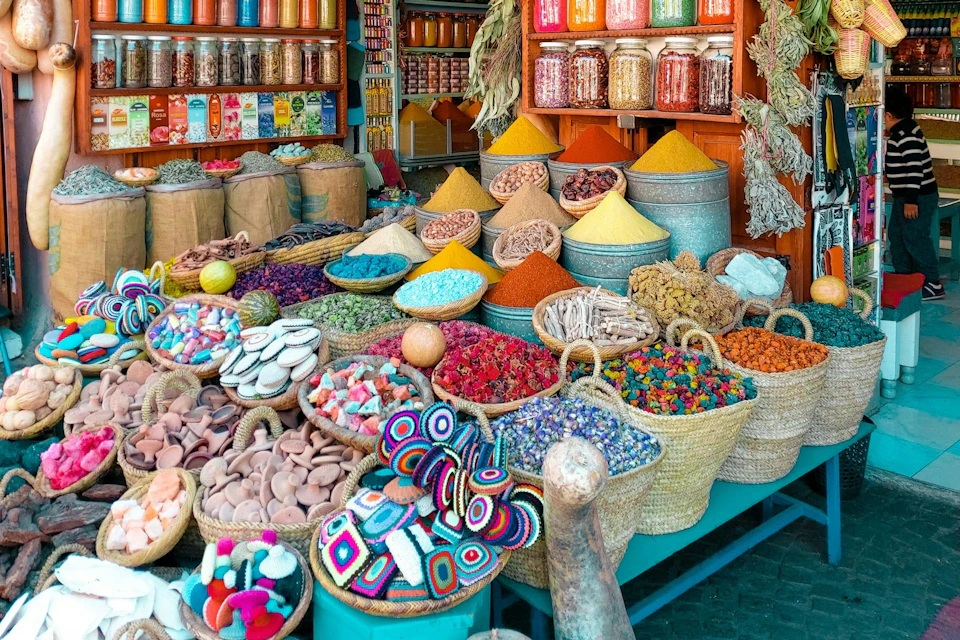 Vibrant market stall showcasing colorful spices, herbs, and dried goods in bowls and baskets, with jars and bags arranged behind.