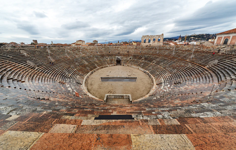 Ancient Roman amphitheater with empty stone seating, an open central arena, and a cloudy sky above.