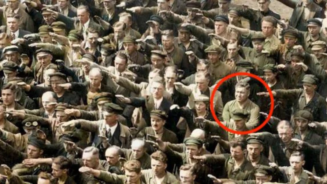 A crowd of people performing a salute, with one person circled in red standing with arms crossed and not participating.