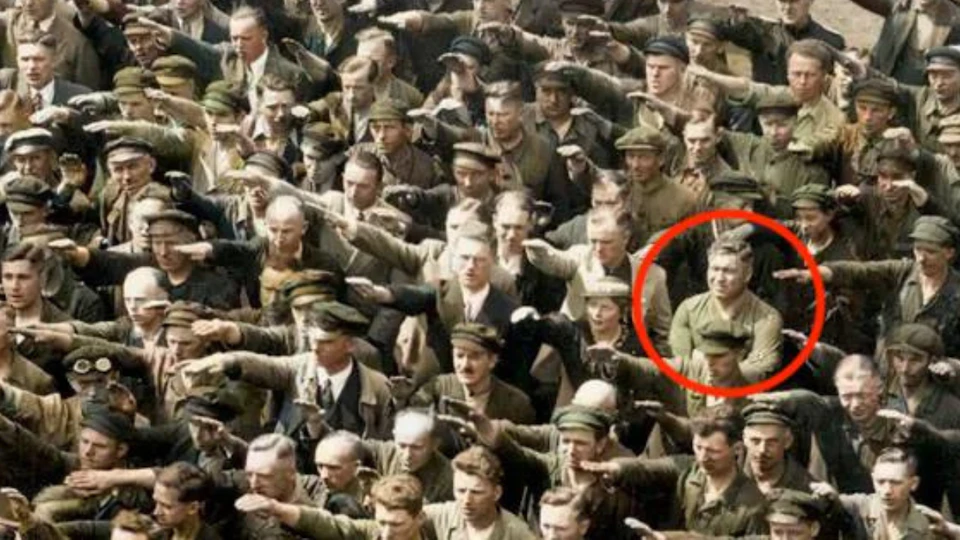 A crowd of people performing a salute, with one person circled in red standing with arms crossed and not participating.