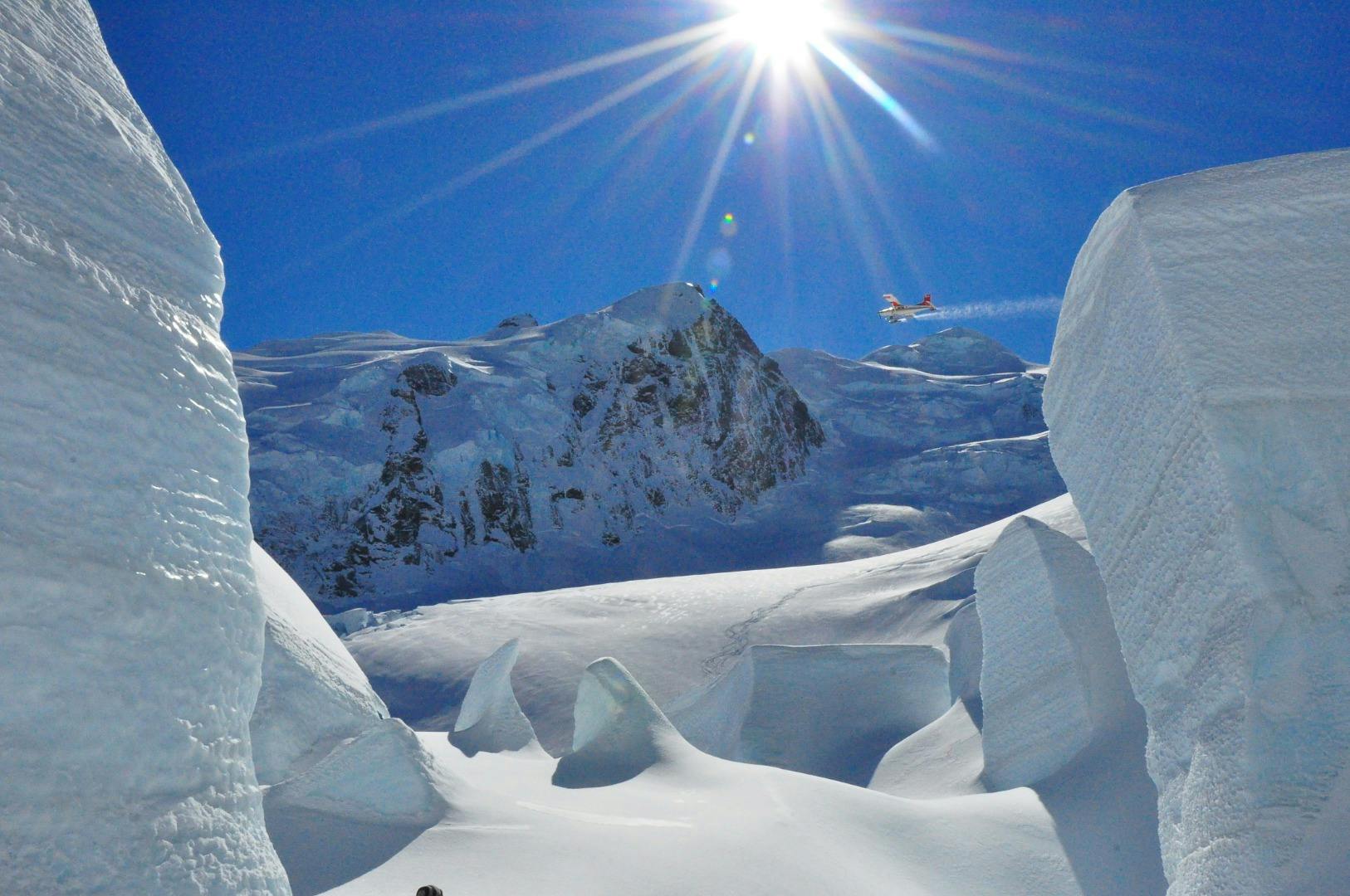 Snowy mountain landscape with steep peaks under a bright sun. A small plane is flying above the scene.