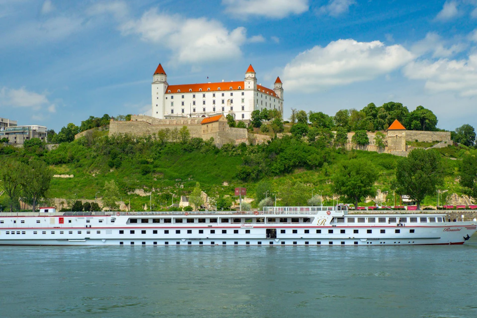 White cruise ship on a river with a large castle featuring four corner towers and red roofs on a green hill under a blue sky.