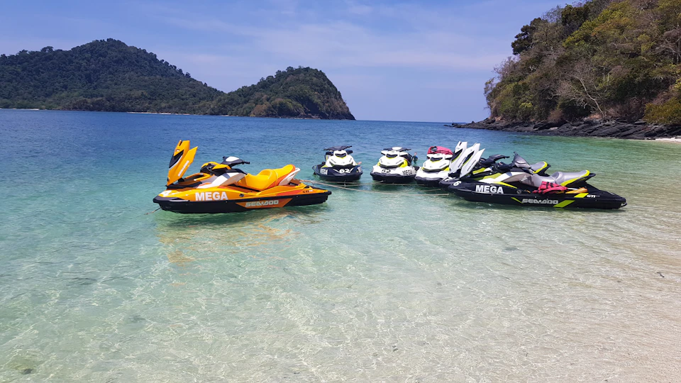 Several jet skis are floating in clear, shallow water near a sandy beach with hilly islands in the background.