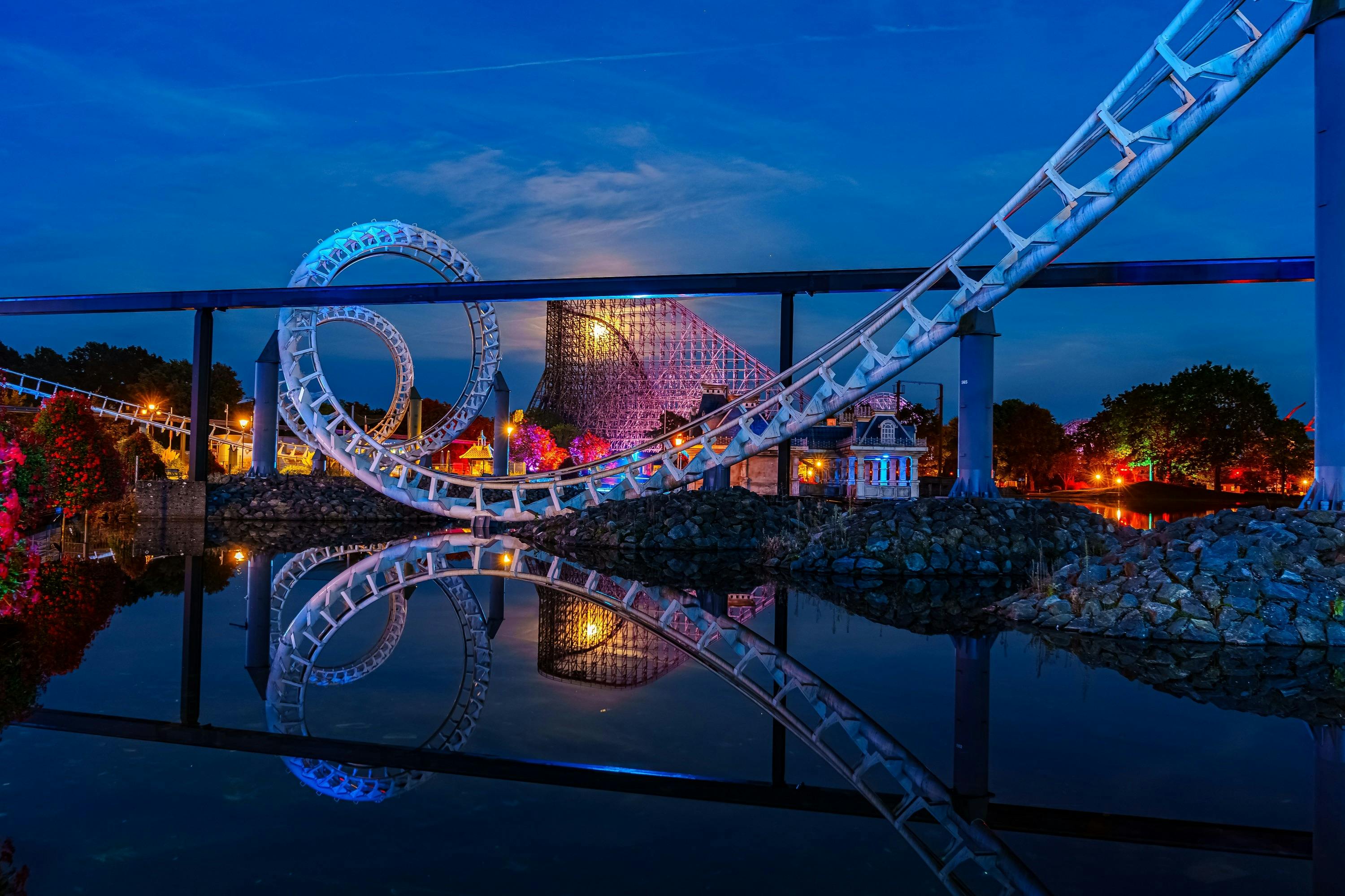 Un ottovolante con loop che si riflettono sull’acqua calma, illuminato di notte, con un ottovolante in legno e un edificio illuminato sullo sfondo.