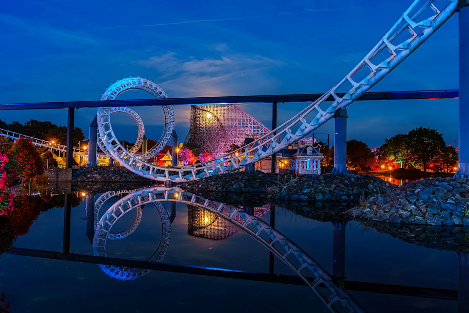 A roller coaster with loops reflected in calm water, illuminated at night with a wooden coaster and a lit building in the background.