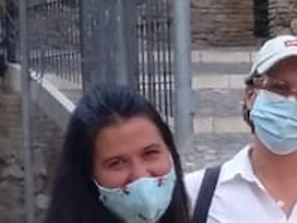 A group of people wearing face masks stands together on a tiled outdoor plaza with trees and historic ruins in the background.
