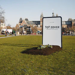 Människor som går i en park med en skylt för "tot ZOVER Nederlands Uitvaart Museum" och historiska byggnader i bakgrunden.