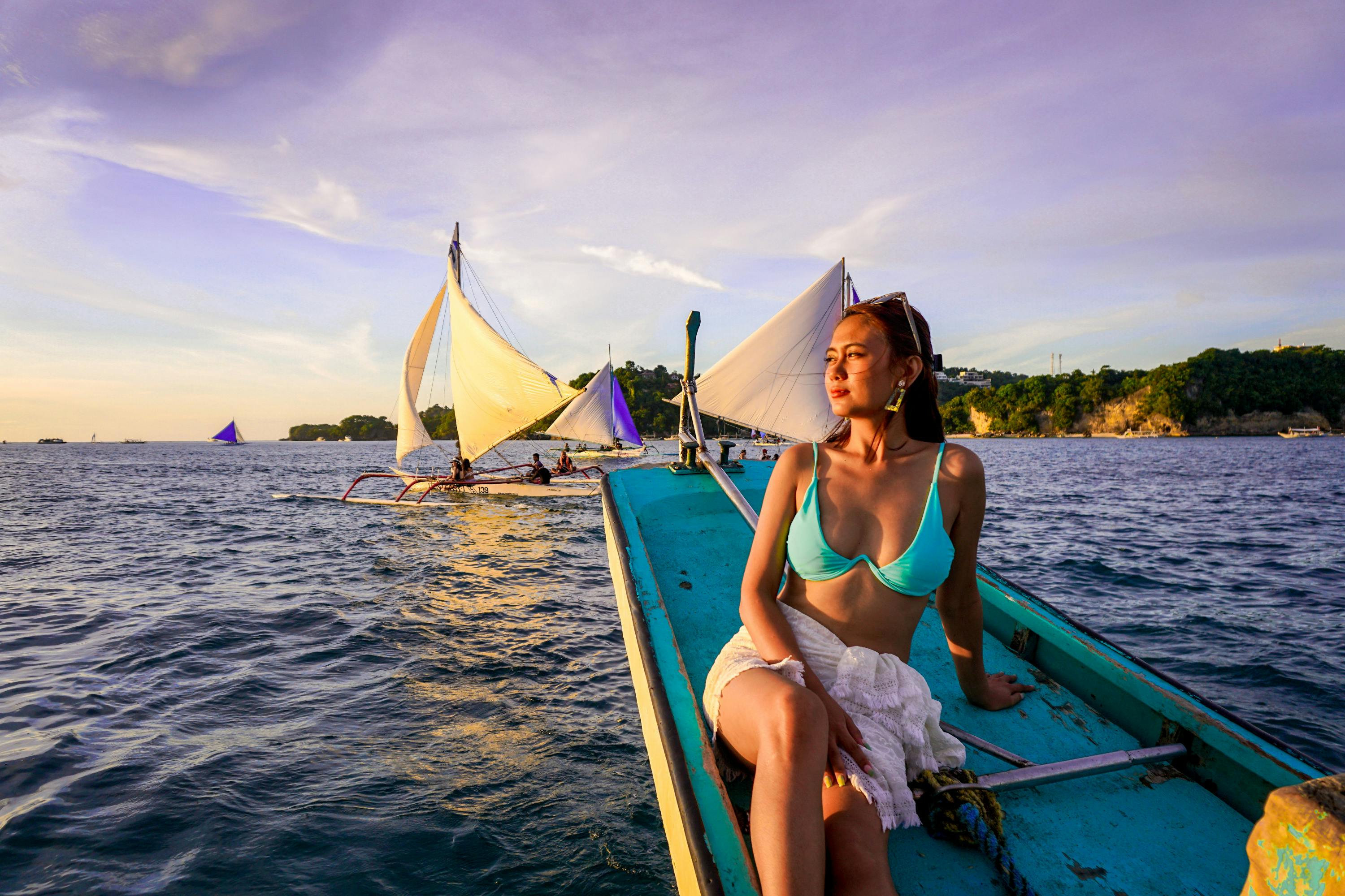 A woman in a turquoise bikini and white wrap sits on the bow of a boat at sunset, with sailboats and a shoreline in the background.