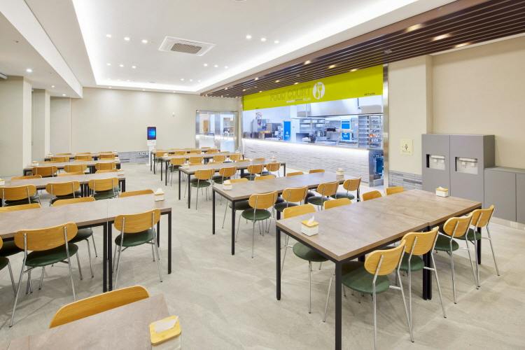 A clean cafeteria with wooden tables and chairs, a service counter in the background, and a bright, spacious interior.