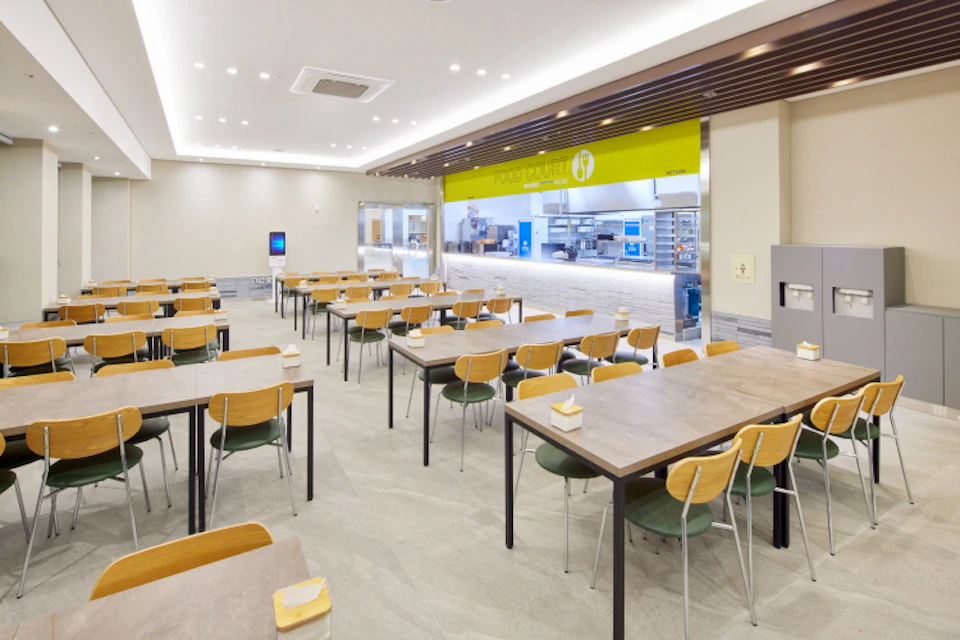 A clean cafeteria with wooden tables and chairs, a service counter in the background, and a bright, spacious interior.