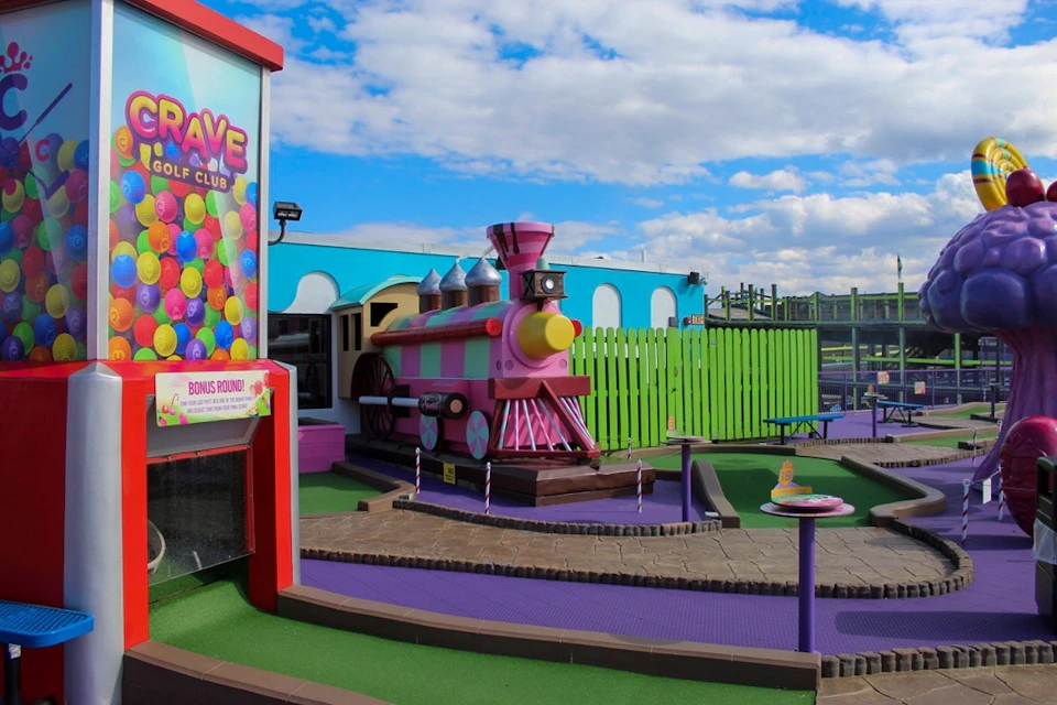 Colorful mini golf course with a pink toy train, bright green fence, and play area under a blue sky with scattered clouds.