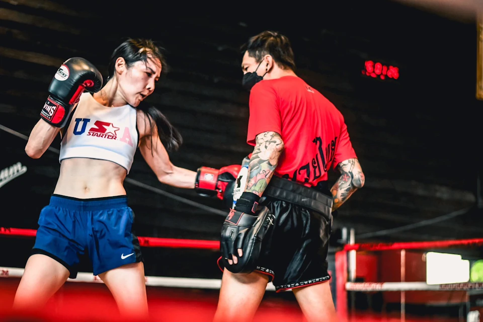 Two boxers sparring in a ring, one wearing blue shorts and a white top, the other in a red shirt and black shorts.