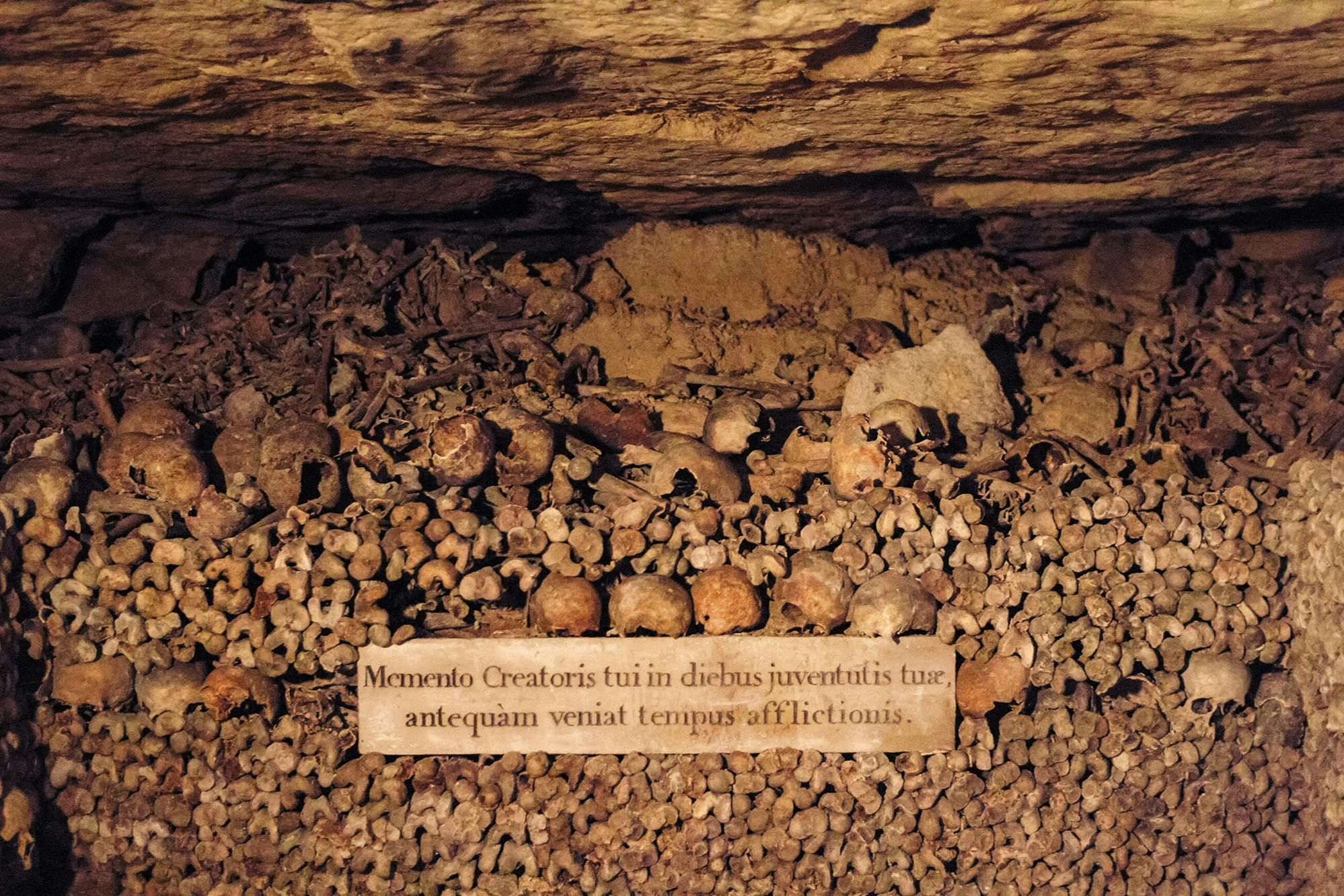 A pile of human bones and skulls arranged in an underground catacomb, with a Latin inscription on a plaque.