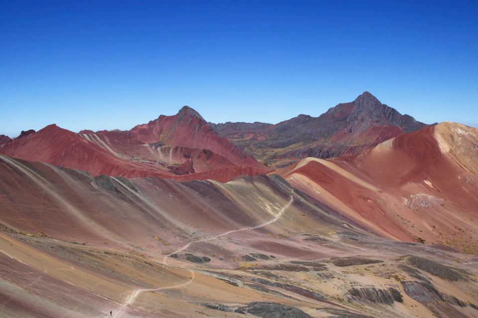 Mountain landscape with vibrant red, yellow, and brown slopes under a clear blue sky, with a winding path through the terrain.
