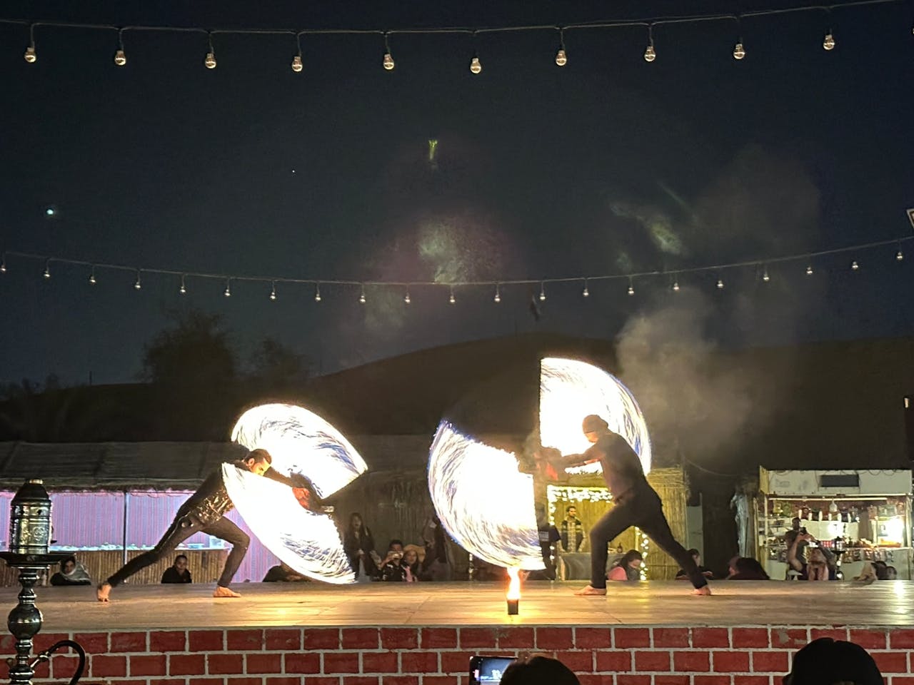 Two performers on a stage, spinning large rings of fire against a dark night sky with string lights above.