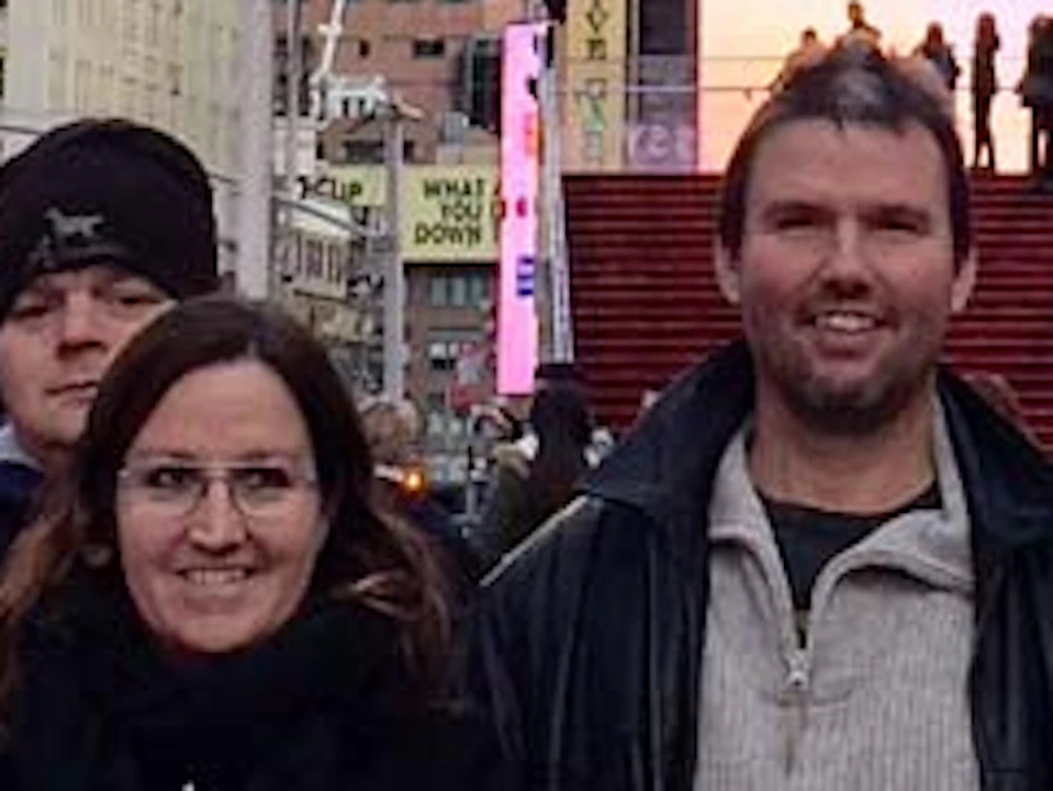 A group of nine people poses in front of a statue of George M. Cohan in Times Square, surrounded by bright advertisements and tall buildings.
